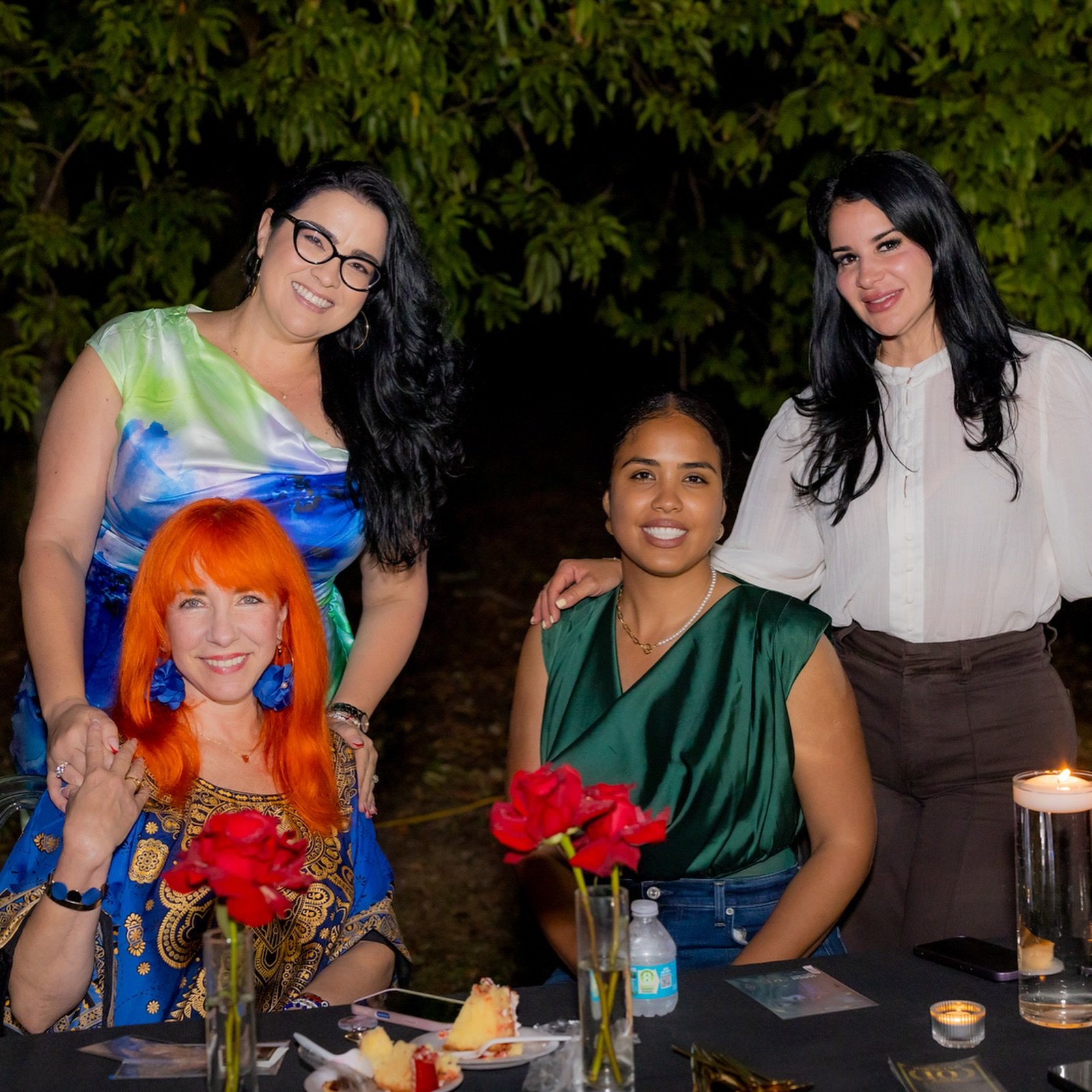 Cuatro mujeres posando al aire libre junto a un arreglo de rosas rojas.